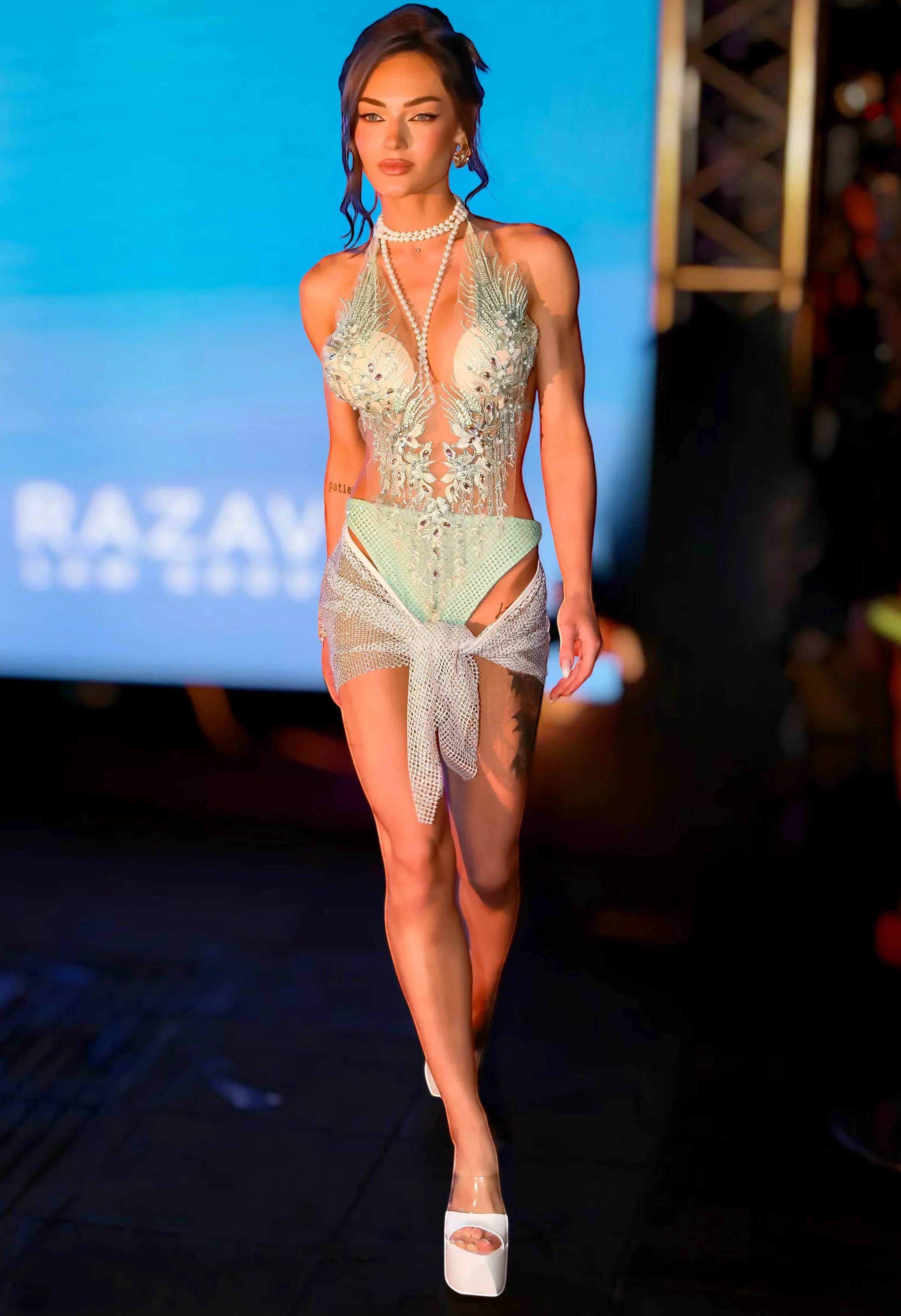 SlaySwimwear mint green one-piece swimsuit with rhinestones, pearls, and feathers. Model wears a sheer skirt.