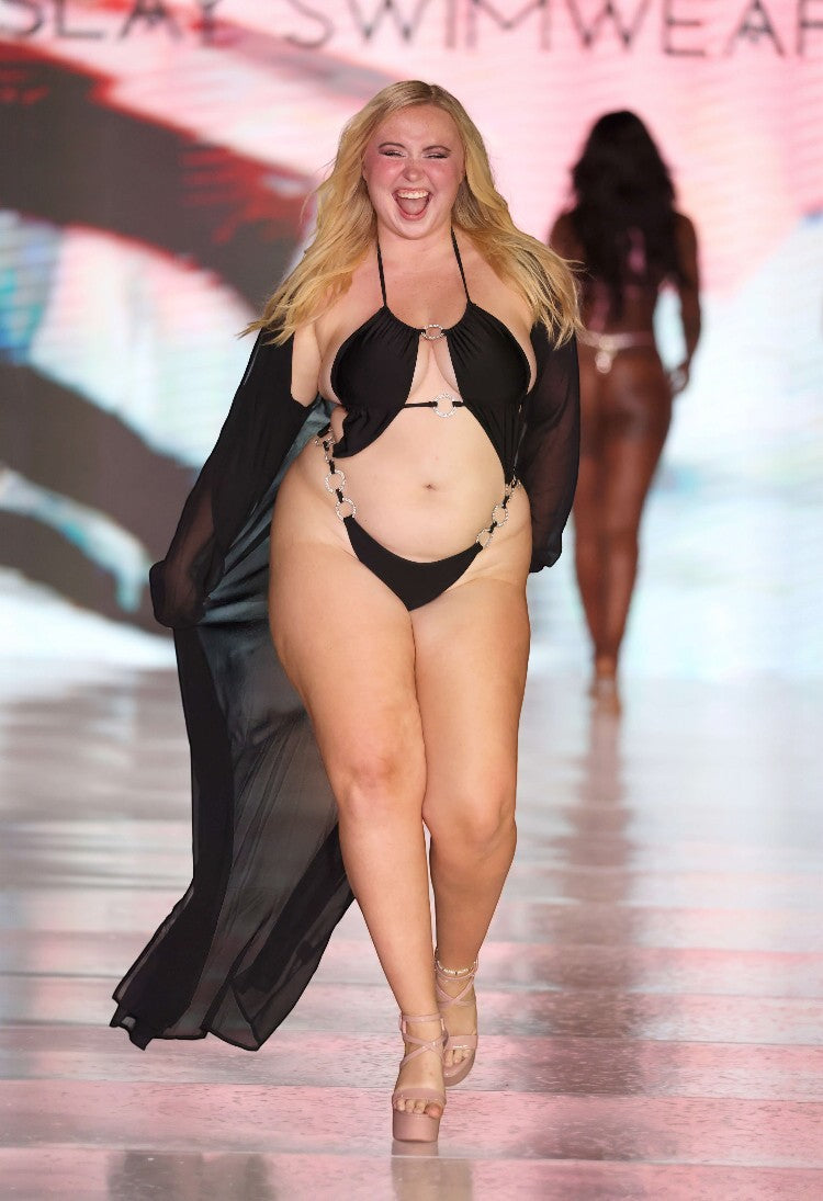 SlaySwimwear black one-piece swimsuit with rhinestone connectors and adjustable straps, worn by a model on a runway.