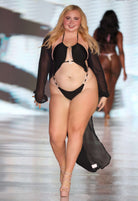 SlaySwimwear black one-piece swimsuit with rhinestone connectors and adjustable straps, worn by a model on a runway.